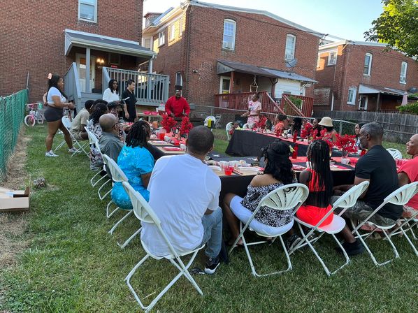 Outdoor family gathering in a brick rowhouse backyard, long lawn dinner with red-and-black table decor and white folding chairs as multigenerational guests enjoy a casual summer evening party