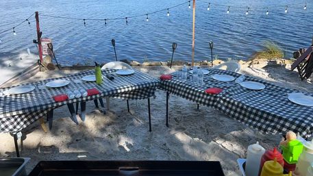 Sandy lakeshore outdoor dining setup with two black-and-white checkered tables, plastic chairs, white plates, bottled water and red napkins under string lights, with grill and condiment bottles in the foreground.