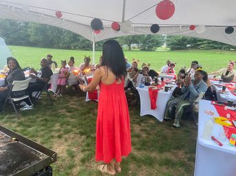 Outdoor family celebration under a white tent on a grassy lawn: woman in a red dress lights handheld torches while guests at red-and-white decorated tables cheer and take photos.