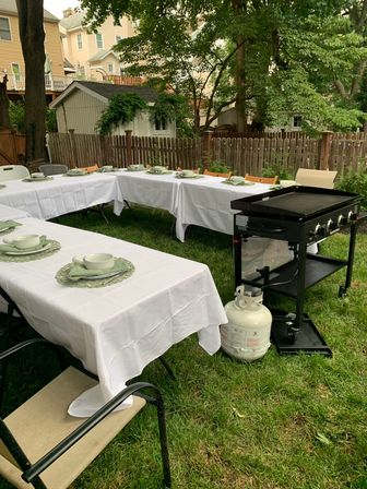 Cozy suburban backyard al fresco setup with U-shaped white-tablecloth tables, green place settings, and a propane griddle on the lawn by a wooden fence and trees.