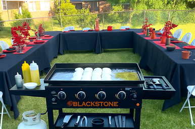 Flat-top griddle in a sunny backyard cooking burger buns, centered inside U-shaped tables draped in black cloths with red place settings and bright red floral accents.