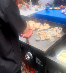 Sizzling outdoor griddle with shrimp, chicken and steak cooking at a casual backyard cookout, guests and drinks on picnic tables in the background.