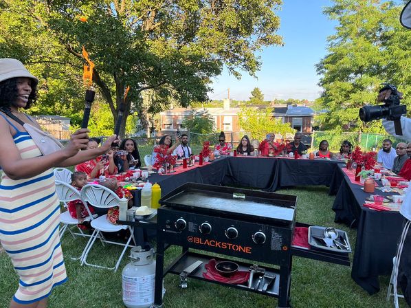 Summer suburban backyard party with U-shaped table and red decor, friends and family seated, a host holding two lit torches by a flat-top grill while a photographer films.