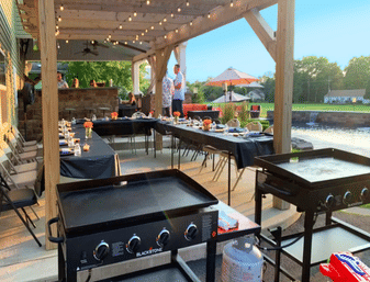 Covered backyard patio dining by a pond with string lights — long tables set for an outdoor dinner with black linens and centerpieces, flat-top griddles in the foreground and umbrella seating in the background.