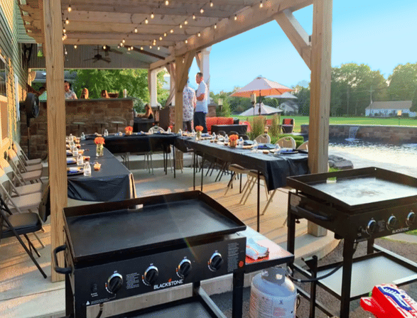 Covered backyard patio dining by a pond with string lights — long tables set for an outdoor dinner with black linens and centerpieces, flat-top griddles in the foreground and umbrella seating in the background.