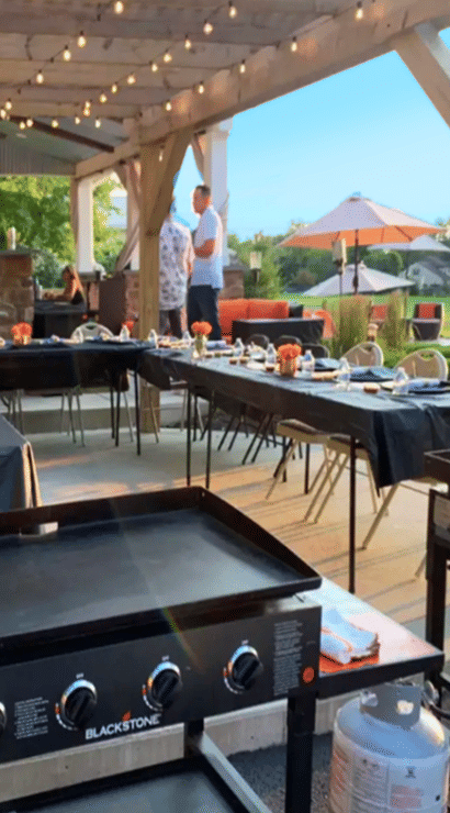 Covered backyard patio dining by a pond with string lights — long tables set for an outdoor dinner with black linens and centerpieces, flat-top griddles in the foreground and umbrella seating in the background.