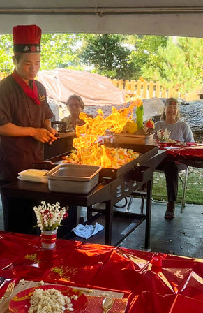 Outdoor backyard teppanyaki chef flambéing vegetables on a flat-top grill with dramatic flames while guests watch at a red-tableclothed party.
