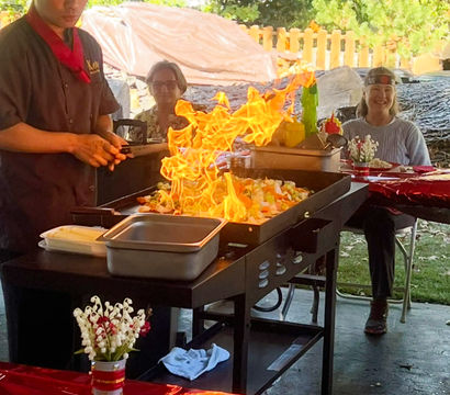 Outdoor backyard teppanyaki chef flambéing vegetables on a flat-top grill with dramatic flames while guests watch at a red-tableclothed party.