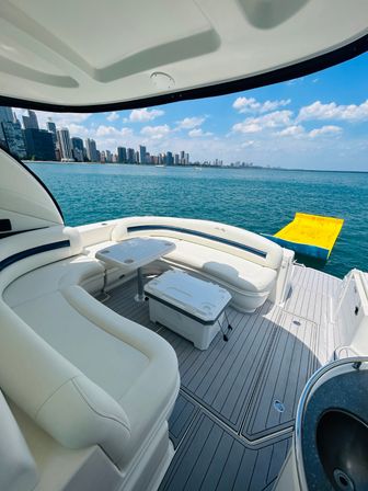 White luxury boat seating and deck with table and cooler, yellow inflatable mat beside, turquoise lake and distant city skyline under a sunny blue sky