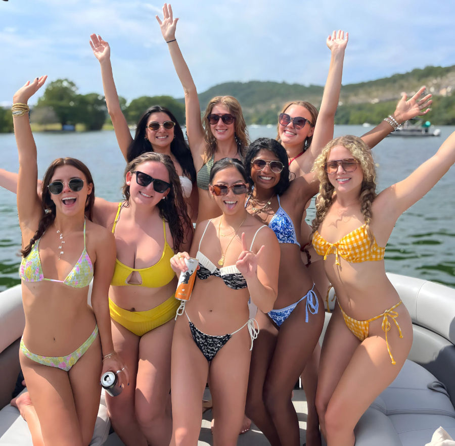 Eight friends in colorful bikinis smiling and cheering on a pontoon boat during a sunny lake day with tree-lined hills in the background
