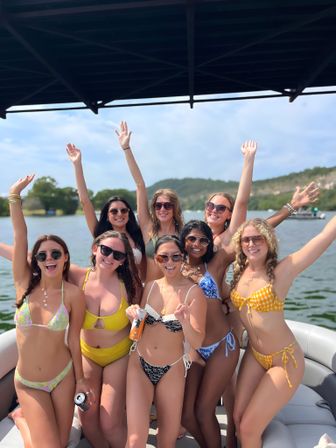 Group of eight friends in colorful bikinis and sunglasses cheering with arms raised on a pontoon boat under a canopy, sunny lake with green hills in the background.