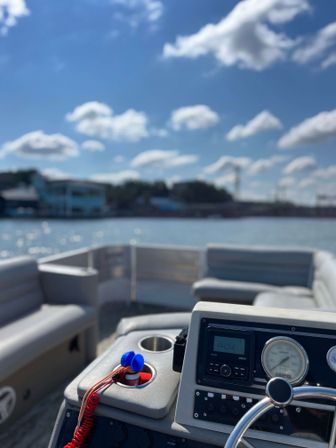 Pontoon boat helm with steering wheel, gauges and cup holder, floating on a sparkling lake under a bright blue sky with fluffy clouds and a distant shoreline.