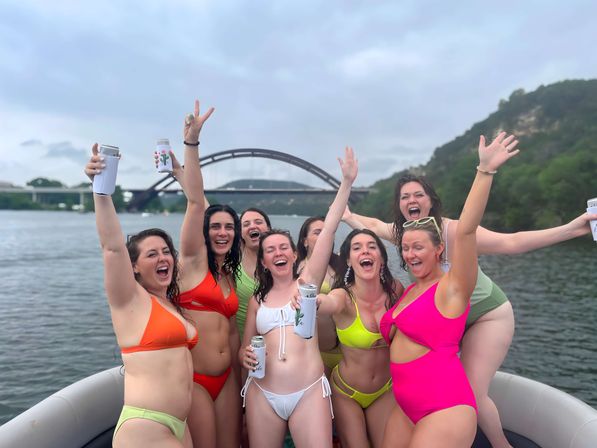 Eight women in colorful swimsuits cheering with drinks on a boat near an arched river bridge and green hills — lively lakeside boat party scene