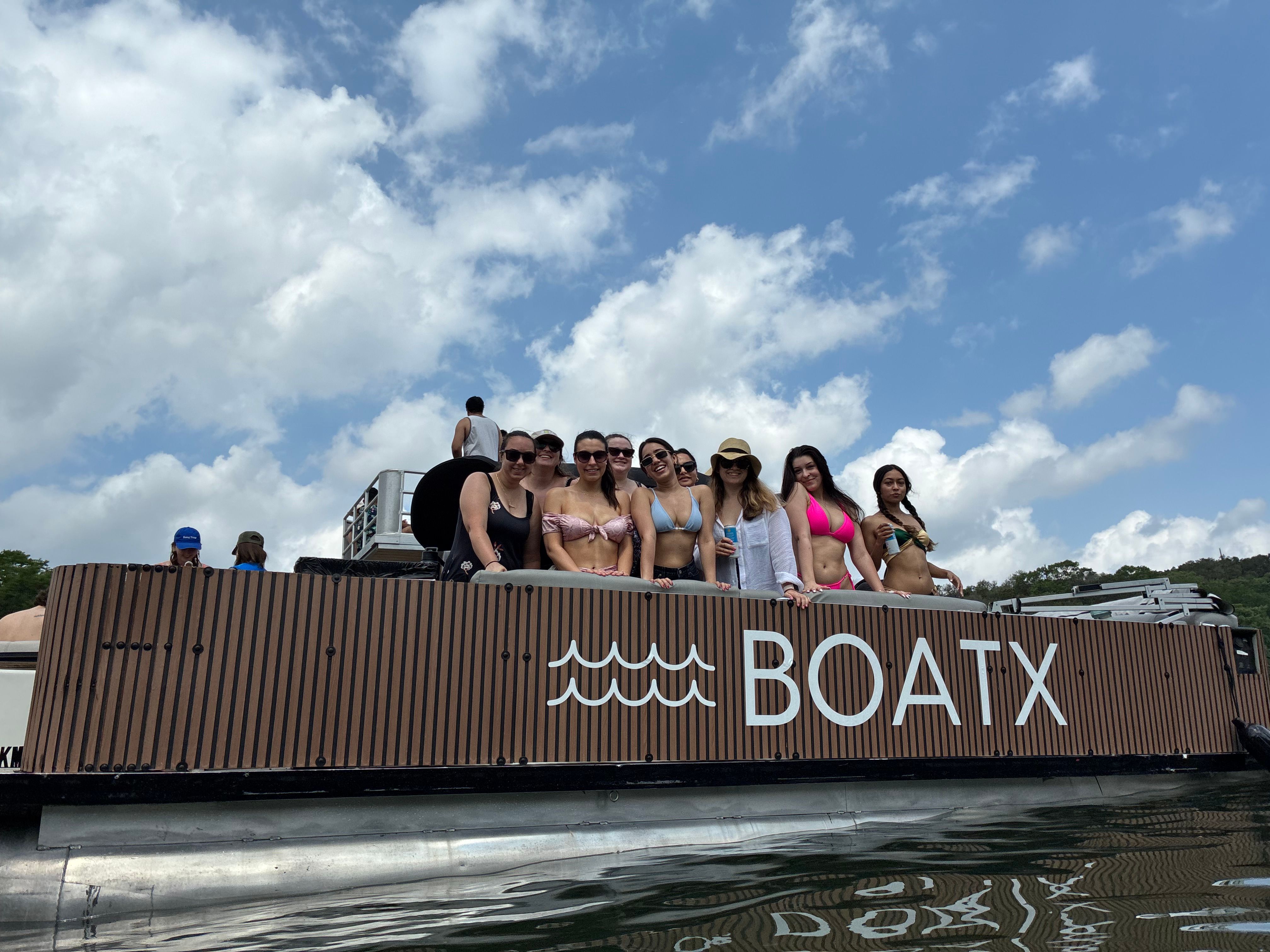 Group of women in swimsuits enjoying a summer lake boat party on a wood-paneled pontoon under a blue sky with puffy clouds.