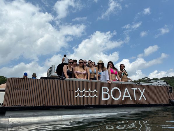 Group of women in swimsuits enjoying a summer lake boat party on a wood-paneled pontoon under a blue sky with puffy clouds.