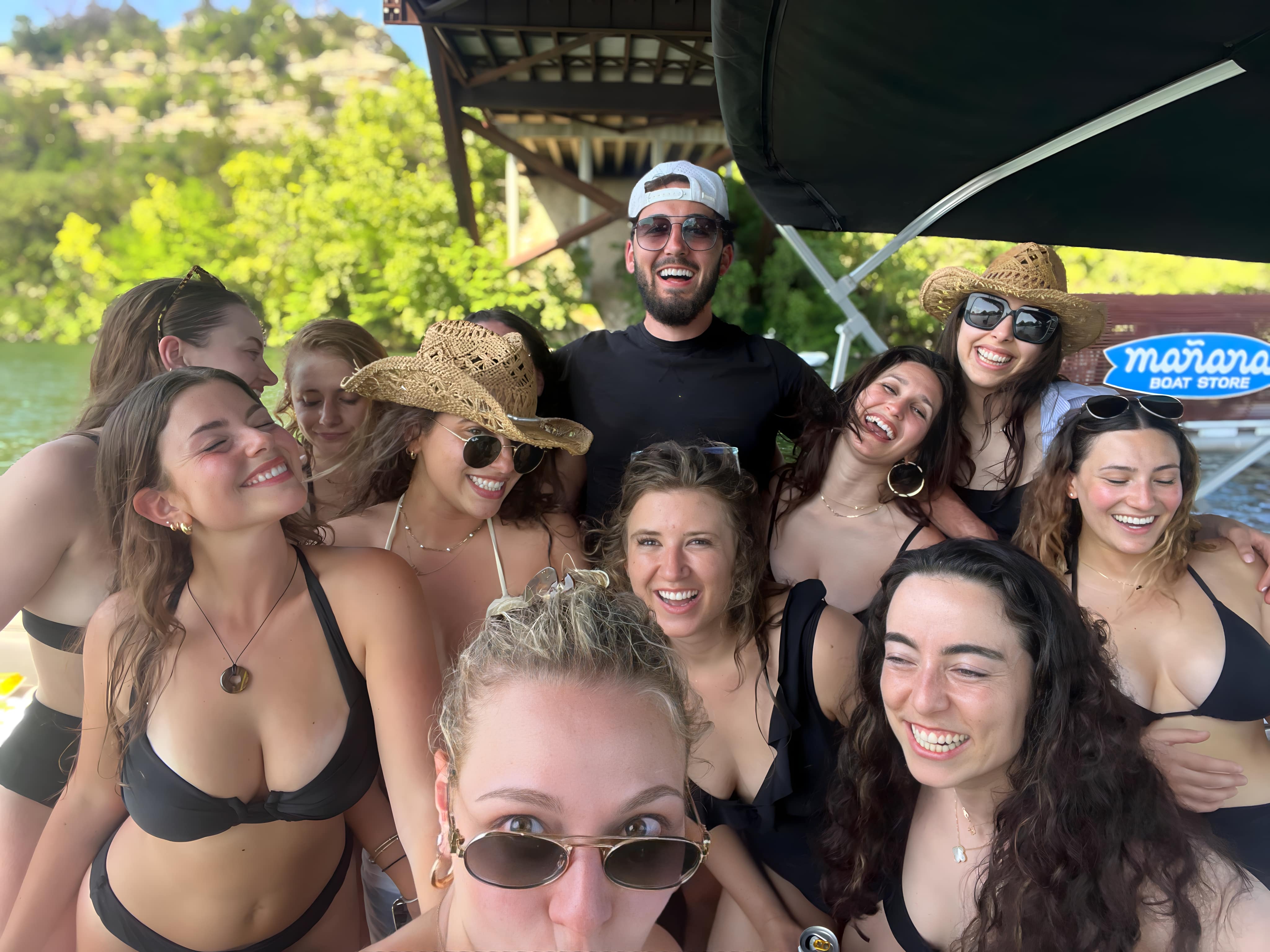 Sunny lake boat selfie of a smiling group in swimsuits, sunglasses and straw hats under a dock with green shoreline — lively summertime boat party vibe.