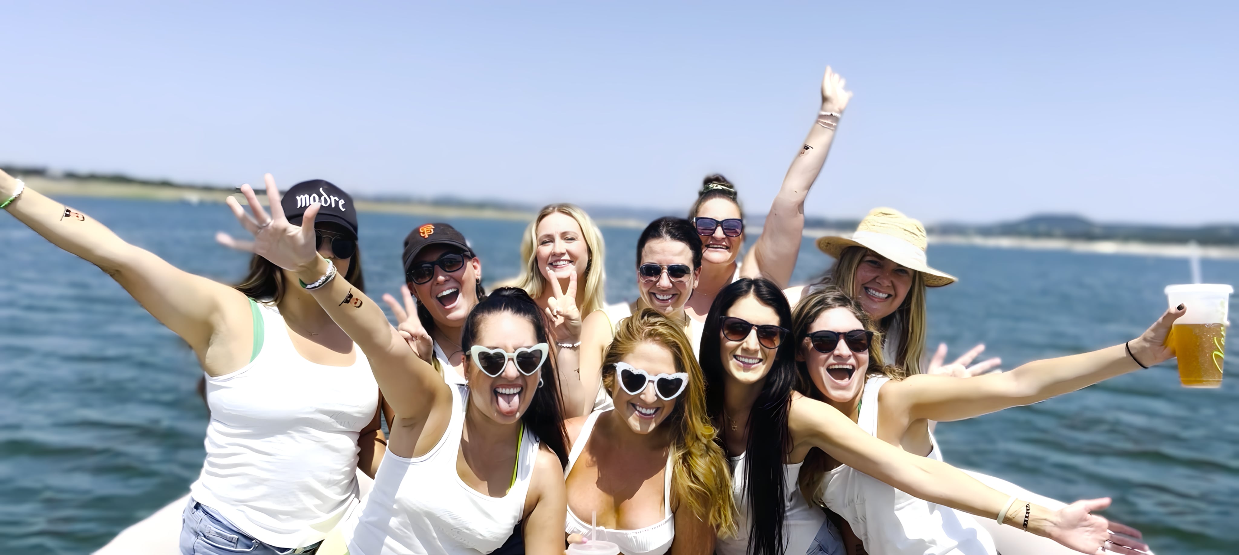Group of friends in white tops and sunglasses cheering on a sunny coastal boat cruise, arms raised and smiling, one holding a beer, ocean and shoreline in background