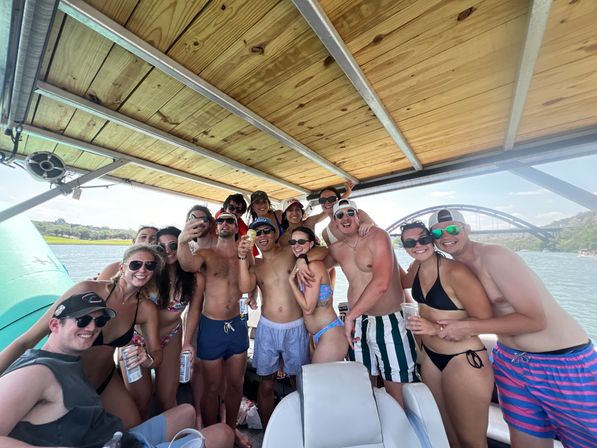 Group of friends smiling and posing on a covered pontoon boat on a sunny lake, wearing swimsuits and sunglasses and holding drinks with an arched bridge visible in the background.