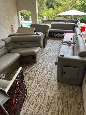Gray vinyl pontoon-style seating with cup holders and speakers on a patterned deck, covered outdoor lounge overlooking a pool and greenery.
