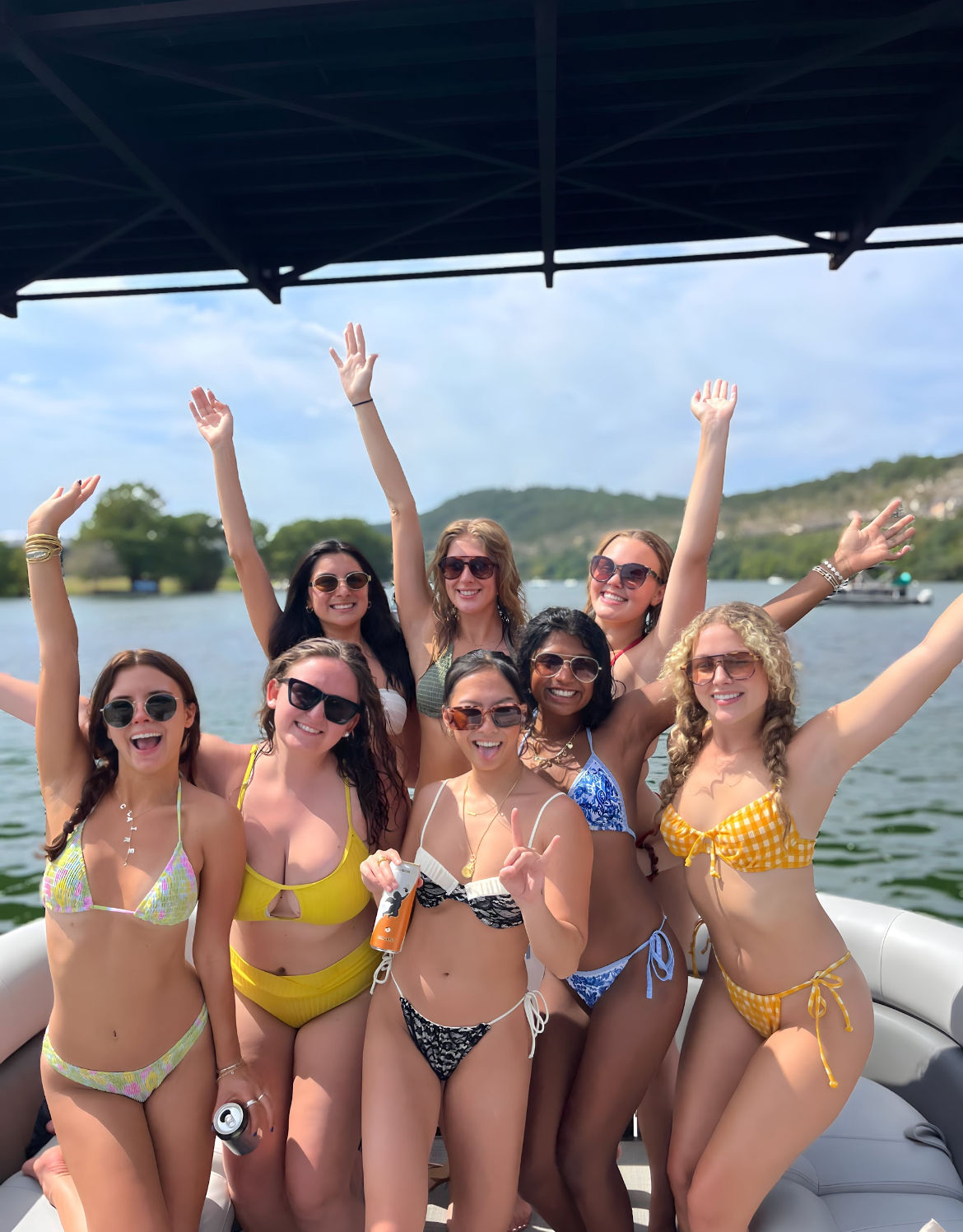 Eight friends in colorful bikinis smiling and raising their arms on a pontoon boat under a canopy on a sunny lake with tree-covered hills in the background