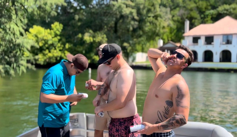 Four men on a motorboat at a sunny lake—two shirtless and one in sunglasses posing with canned drinks, trees and a waterfront house visible in the background.