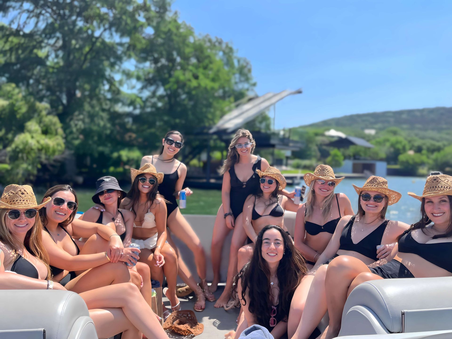 Group of friends in swimsuits and straw cowboy hats smiling and relaxing on a pontoon boat on a sunny lake with green trees and hills in the background.