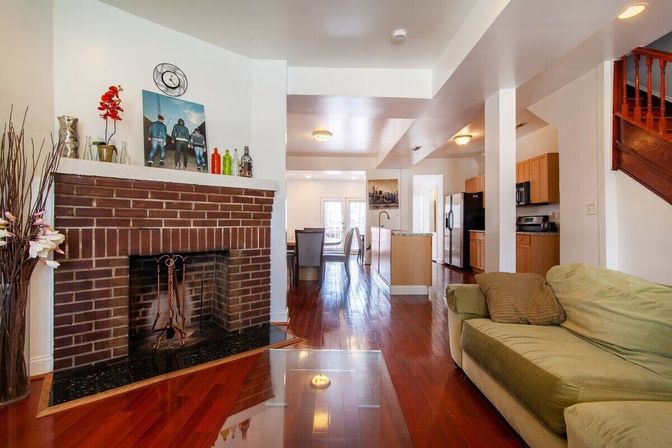 Cozy open-concept living room in a townhouse with a brick fireplace, glossy hardwood floors, green sofa, and a modern kitchen with stainless-steel appliances.