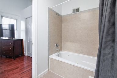 Bright apartment bathroom with beige tiled bathtub-shower combo, chrome fixtures and gray shower curtain, partial view into adjacent bedroom with dark wood dresser, flat-screen TV and cherry hardwood floors.