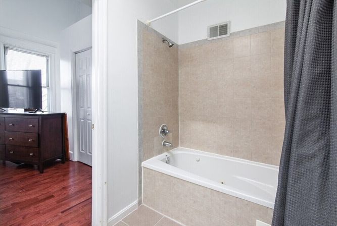 Bright apartment bathroom with beige tiled bathtub-shower combo, chrome fixtures and gray shower curtain, partial view into adjacent bedroom with dark wood dresser, flat-screen TV and cherry hardwood floors.