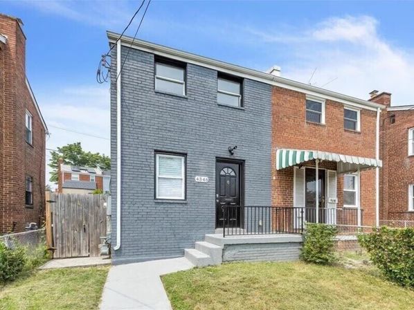 Two-story urban brick rowhouse painted gray attached to a red-brick neighbor, featuring a small front porch with black door and railing, green-and-white striped awning, front yard and sidewalk under a blue sky.