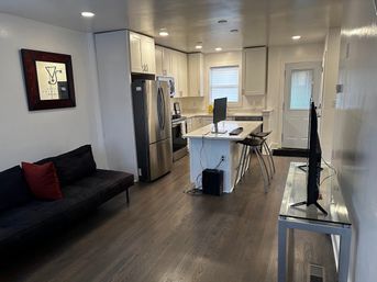 Cozy modern open-concept apartment kitchen and living area with white cabinets, stainless-steel refrigerator, central island with computer monitor and metal bar stools, dark hardwood floors, black sofa with red pillow, wall art and TV on a glass console.