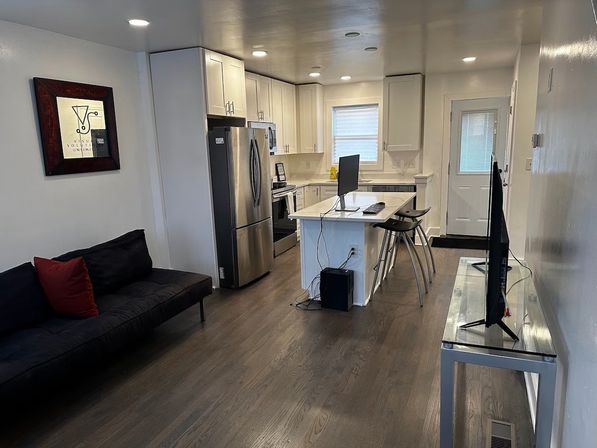 Cozy modern open-concept apartment kitchen and living area with white cabinets, stainless-steel refrigerator, central island with computer monitor and metal bar stools, dark hardwood floors, black sofa with red pillow, wall art and TV on a glass console.