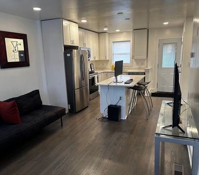 Cozy modern open-concept apartment kitchen and living area with white cabinets, stainless-steel refrigerator, central island with computer monitor and metal bar stools, dark hardwood floors, black sofa with red pillow, wall art and TV on a glass console.