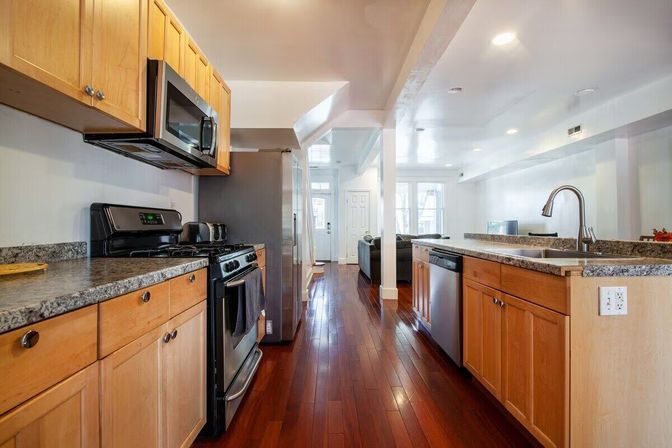 Bright open-concept kitchen with light wood cabinets, granite countertops, stainless steel appliances, center island sink and glossy hardwood floors leading to a living area.