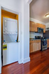Stacked washer-dryer in a small closet beside a modern kitchen with maple cabinets, granite countertops, stainless-steel stove and microwave, and glossy hardwood floors.