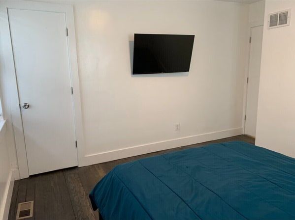 Minimal modern bedroom with teal comforter, wall-mounted flat-screen TV on white walls, hardwood floors, baseboard trim and two doors.