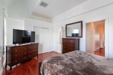 Cozy modern master bedroom with white walls, cherry hardwood floors, dark wood dresser and tall chest with mirror, plush brown bedspread and flat-screen TV