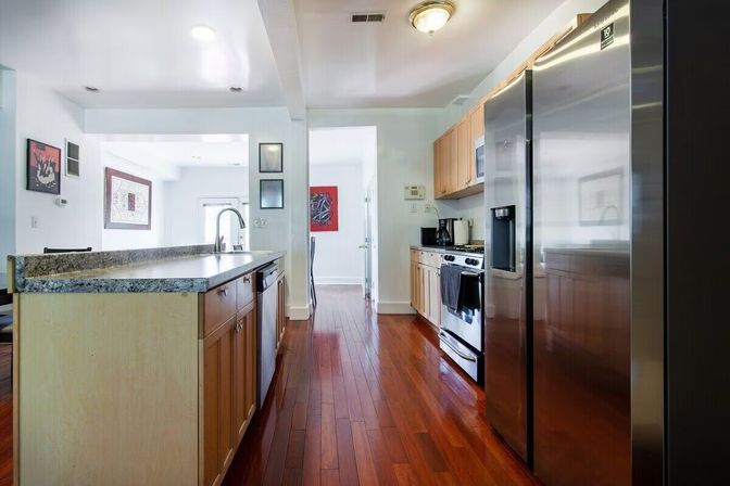 Sleek modern open-concept kitchen with granite-topped island and sink, light wood cabinets, stainless steel refrigerator and oven, microwave, and polished hardwood floors.