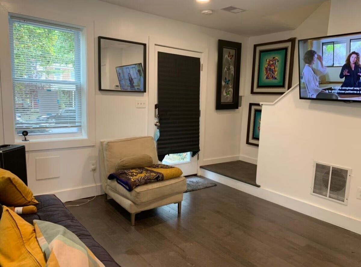 Cozy modern apartment living room with hardwood floors, beige lounge chair with patterned throw, navy sofa with mustard pillows, wall-mounted TV, framed artwork, window blinds and a black door shade.