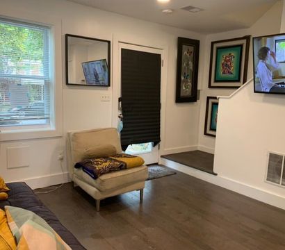 Cozy modern apartment living room with hardwood floors, beige lounge chair with patterned throw, navy sofa with mustard pillows, wall-mounted TV, framed artwork, window blinds and a black door shade.