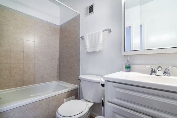 Bright, tidy compact bathroom with a white vanity and chrome faucet, medicine cabinet mirror, white toilet, and beige tiled bathtub-shower with towel bar — modern neutral design for an apartment or home.