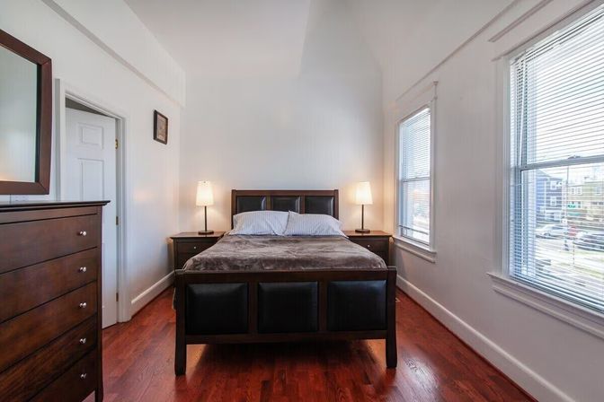 Cozy sunlit city apartment bedroom with dark wood bed, matching dresser and nightstands, two bedside lamps, large blinds-covered windows and warm hardwood floors