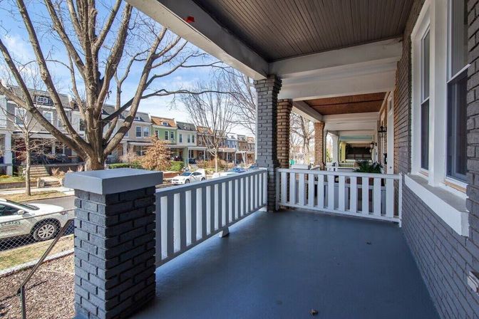 Front porch with dark-painted brick columns and white railing overlooking a tree-lined residential street of rowhouses and parked cars.