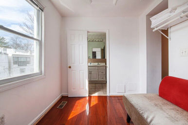 Sunlit small apartment room with glossy hardwood floors, white walls, large window overlooking nearby buildings, red upholstered bench and open door to tiled en-suite bathroom with vanity