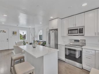 Airy open-concept modern white kitchen with island and white countertop, stainless-steel refrigerator, range and microwave, bar stools, hardwood floors and a small living area with sofa visible in the background.