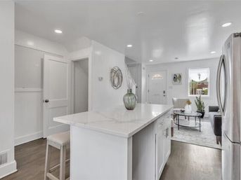 Bright modern white kitchen with marble-topped island and stool, glass vase centerpiece, stainless steel refrigerator and open living room with sofa and round coffee table on hardwood floors.