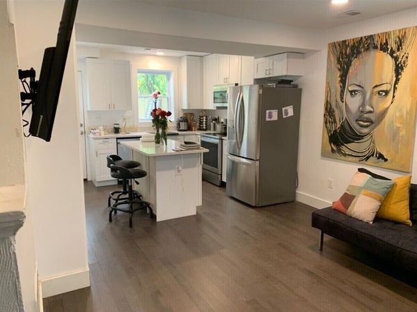 Modern open-concept kitchen and living area with white cabinets, central island and black bar stools, stainless steel fridge, dark hardwood floors, vase of flowers and large portrait artwork on the wall