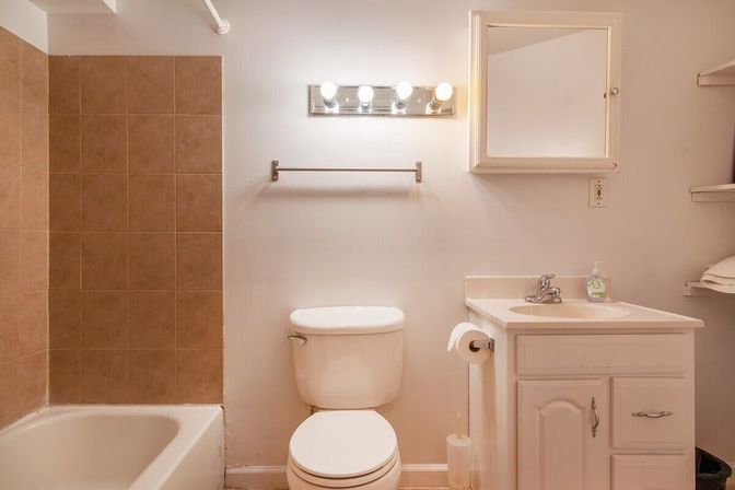 Cozy residential bathroom with white toilet and vanity, bathtub with brown tile surround, mirrored medicine cabinet and four-bulb vanity light