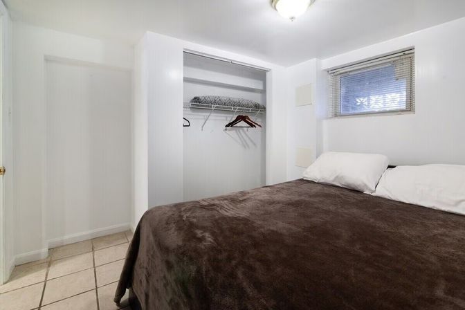 Small basement-style bedroom with high window, white walls, tiled floor, open closet with hangers, and a bed with brown blanket and white pillows