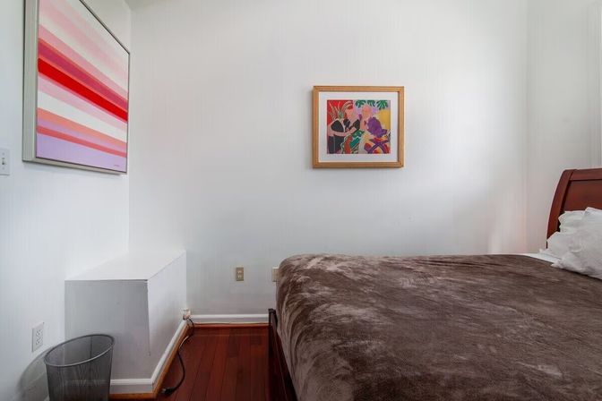 Minimalist bedroom with white walls and mahogany hardwood floor, wooden bed covered in a brown plush blanket, two colorful framed prints on the wall, small white shelf and a metal wastebasket by an electrical outlet.